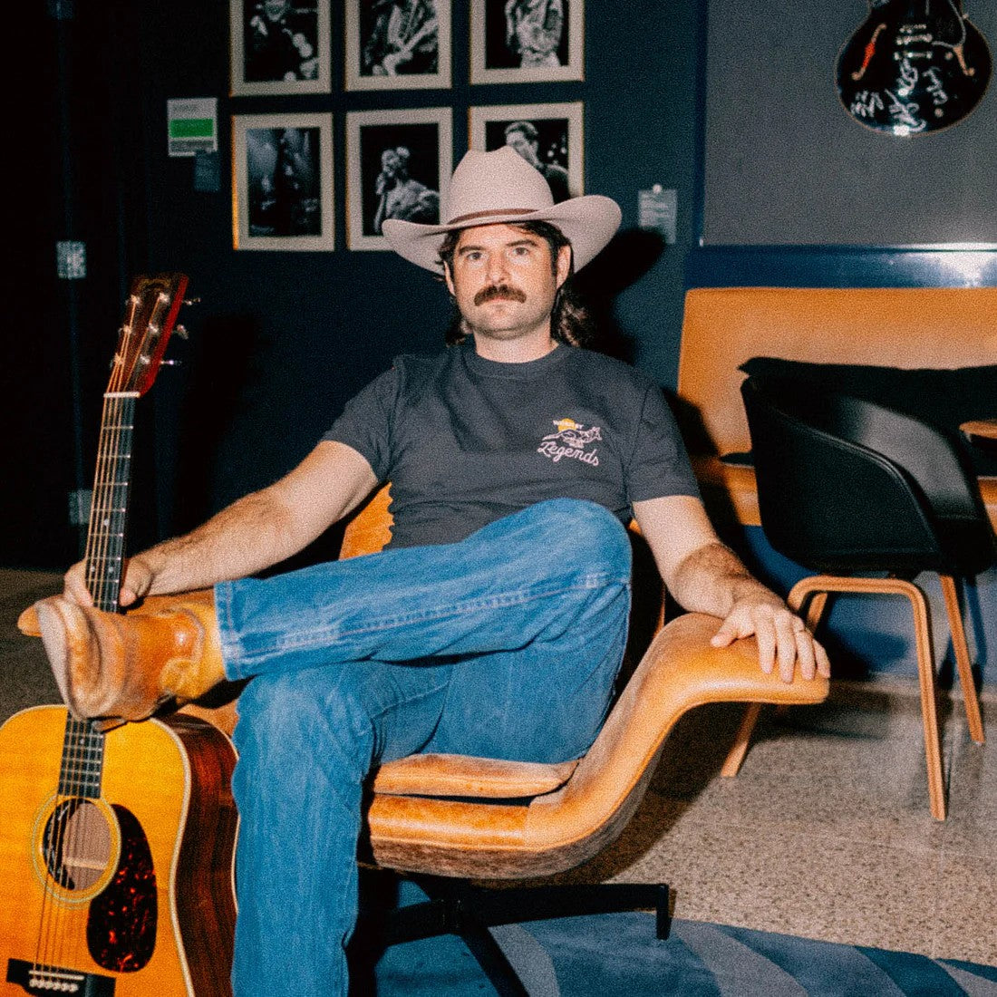 Man in cowboy hat and jeans sitting on a wooden chair holding an acoustic guitar in a room with framed pictures on the wall.