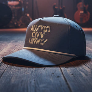 A denim blue trucker-style cap with 'AUSTIN CITY LIMITS' embroidered in cream on the front, resting on a dark wooden surface against a beige background.