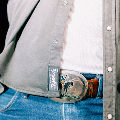 Close-up of a belt with a horse design and a brand label on a person wearing a gray shirt and blue jeans.