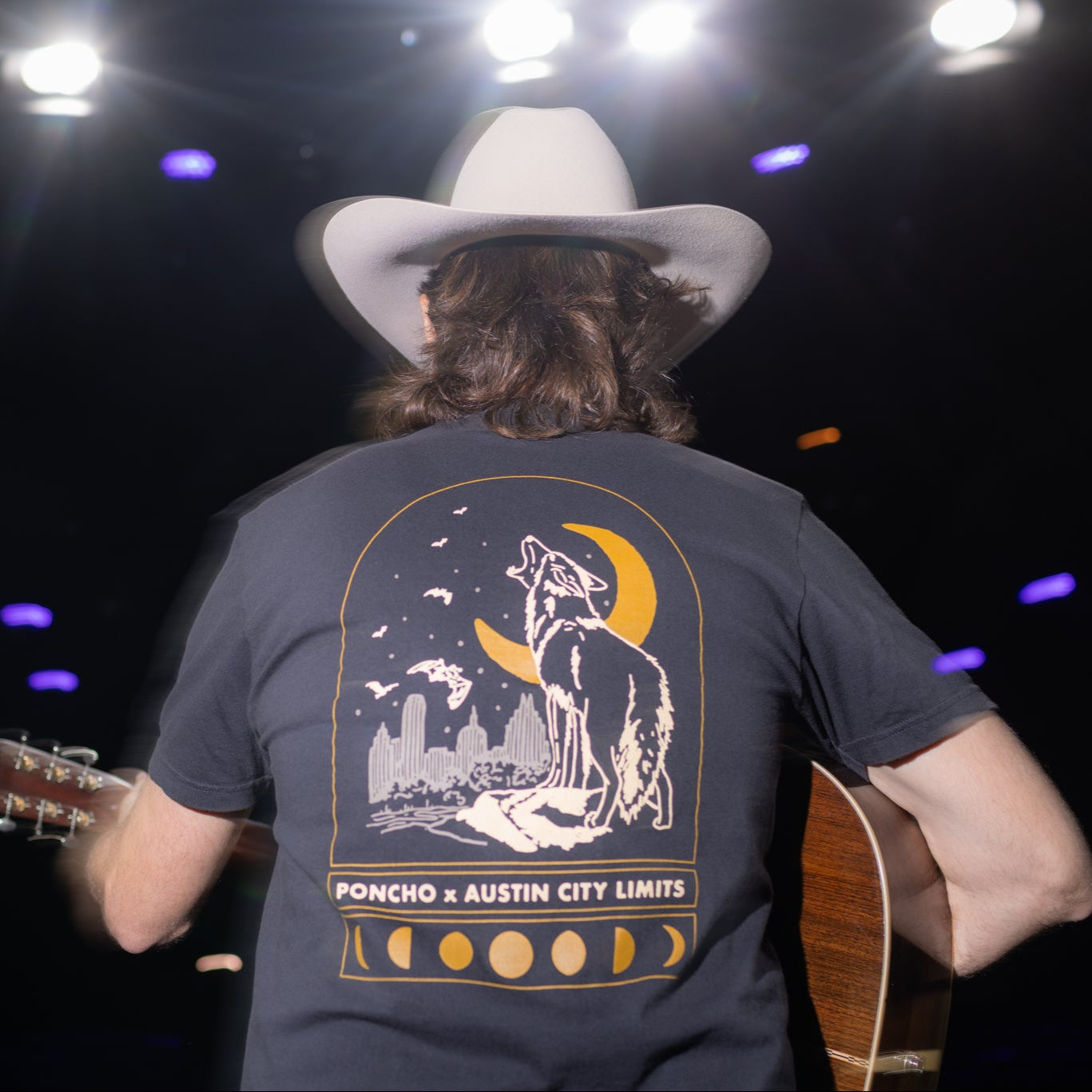 Person on stage wearing a cowboy hat and a t-shirt with a graphic design, holding an acoustic guitar.