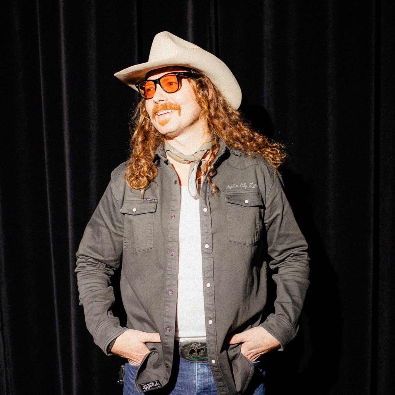 Person wearing a cowboy hat and sunglasses against a black curtain