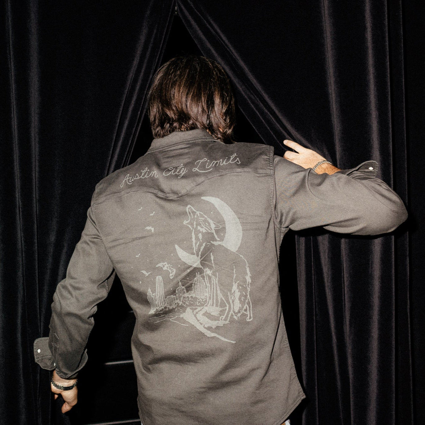 Person wearing a jacket with a design and text on the back, standing in front of black curtains.