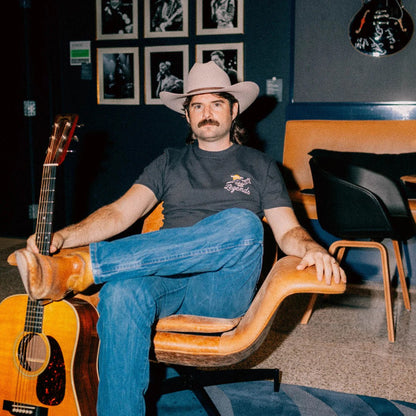 Man in cowboy hat and jeans sitting on a wooden chair holding an acoustic guitar in a room with framed pictures on the wall.